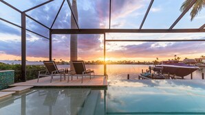 Una piscina infinita con vistas a la puesta de sol sobre el agua, sillones de descanso y un muelle con barcos.