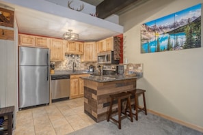 A kitchen with wooden cabinets, stainless steel appliances, a granite countertop, and a painting of a mountain lake.