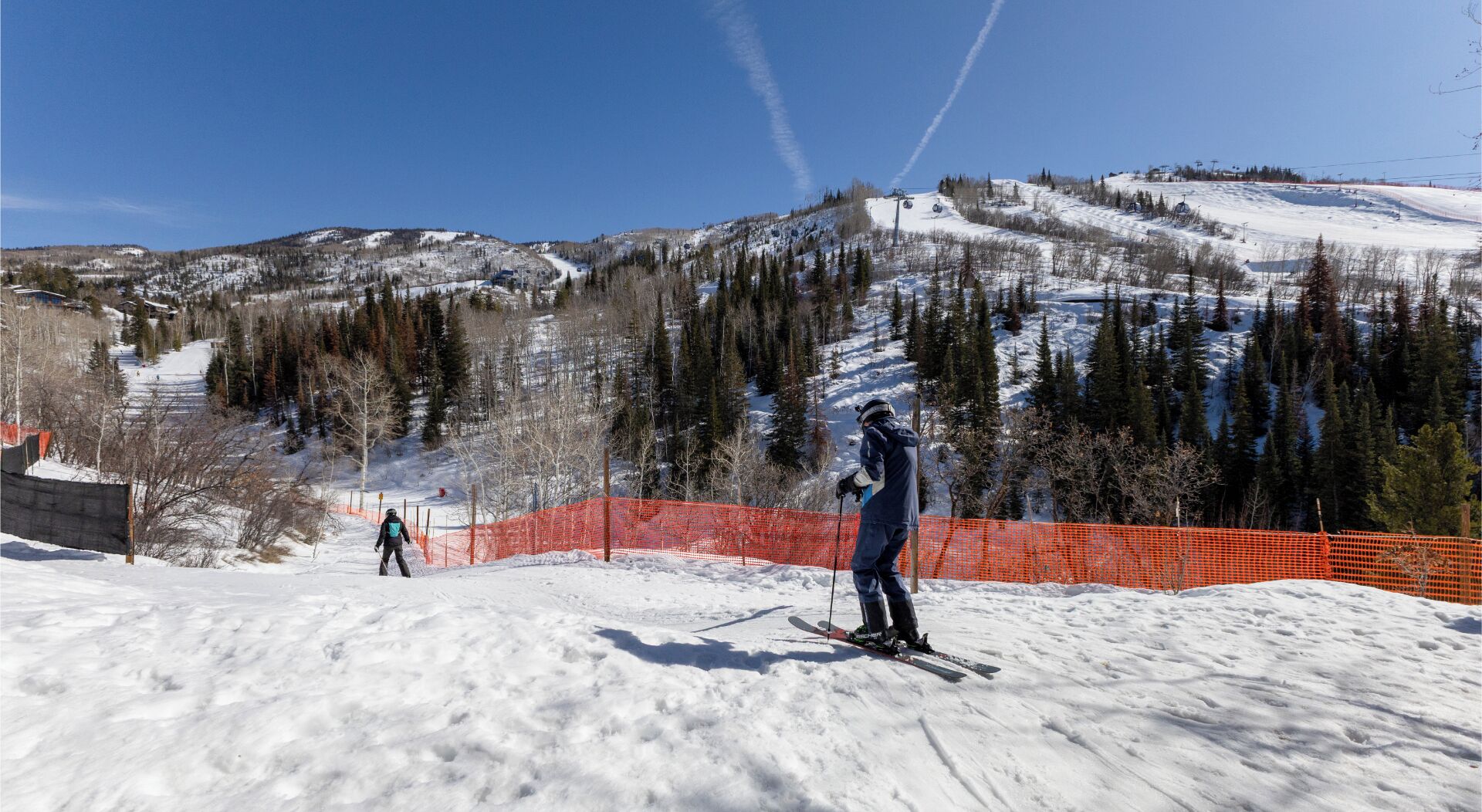 Une piste de ski sous un ciel dégagé, un skieur au premier plan et une montagne boisée en arrière-plan.