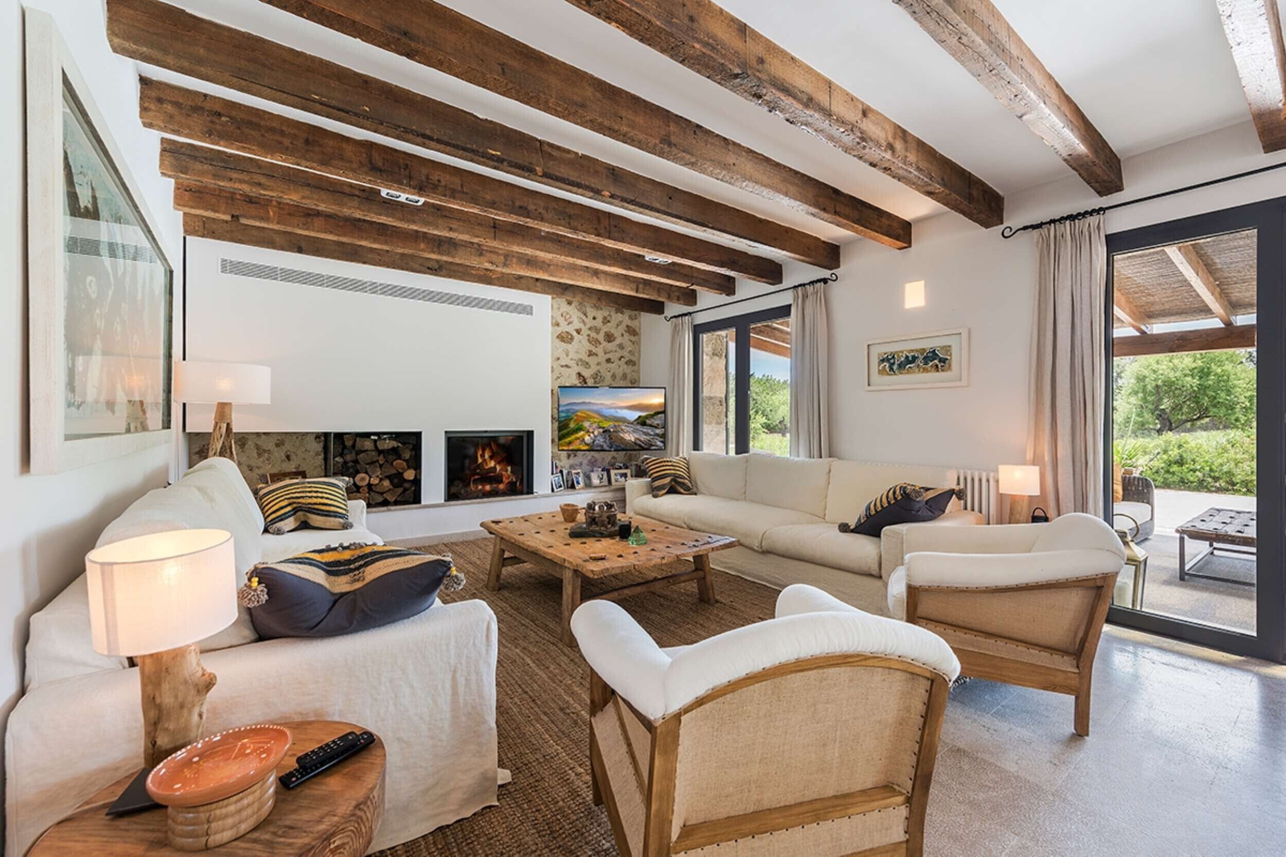 A cozy living room with a fireplace, wooden beams, a sofa set, and a large window with a view of greenery.