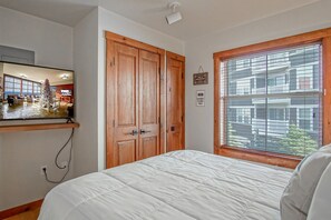 Een slaapkamer met een bed, een houten kledingkast, een raam met zonwering en een televisie aan de muur.
