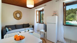 A compact living space with a sofa, dining table, and a refrigerator. A bowl of oranges is on the table, and there's a view of a forested area through a window. - Dina house (Lakka)