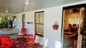 A room with a red chair, a small table, and a hammock. There is a large window and a door leading to another room. - The Flowers House - Downtown Historic Home in the Heart of Mount Dora (Mount Dora)