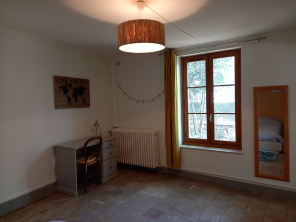 Ett rum med ett element, ett skrivbord med en stol, en hängande lampa, ett fönster med gardiner och en säng med en sänggavel.