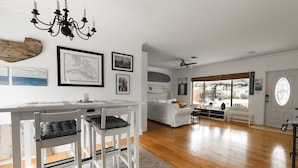 Amplia sala de estar con una mesa de comedor blanca, sillas y una araña. Se observan cuadros en la pared, un sofá blanco y tablas de surf al fondo.