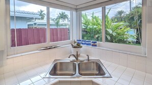 Una cocina con un lavamanos doble, salpicadero de azulejos blancos y amplias ventanas que ofrecen vistas a una cerca roja y follaje exterior.