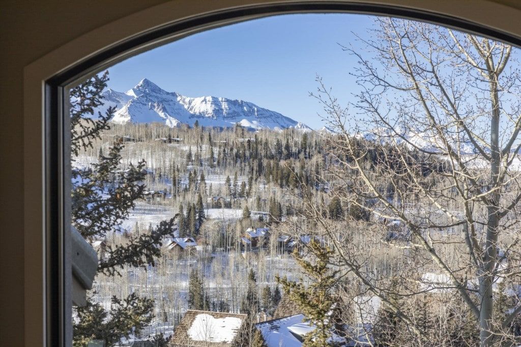 Vista pela janela de uma paisagem montanhosa com picos nevados, área arborizada e alguns edifícios.