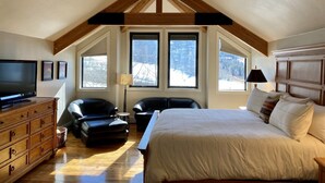 A bedroom with a large bed, a television, a desk, and a chair. There are windows with a view of trees and a ceiling fan. - 226 Country Club Dr 5 Bedroom House (Mountain Village)