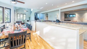 A spacious kitchen with a central island, large windows, and a dining area with chairs and a table set for a meal. - 226 Country Club Dr 5 Bedroom House (Mountain Village)