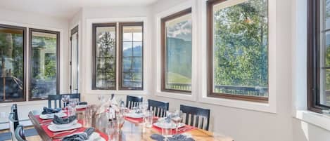 Un comedor con una mesa grande dispuesta para seis personas, rodeado de ventanas que ofrecen vistas a la vegetación y colinas.