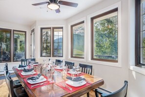 Un comedor con una mesa grande dispuesta para seis personas, rodeado de ventanas que ofrecen vistas a la vegetación y colinas.