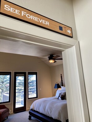 A bedroom with a bed, a ceiling fan, a nightstand, and a lamp. There is a large window and a door leading to an outdoor area.