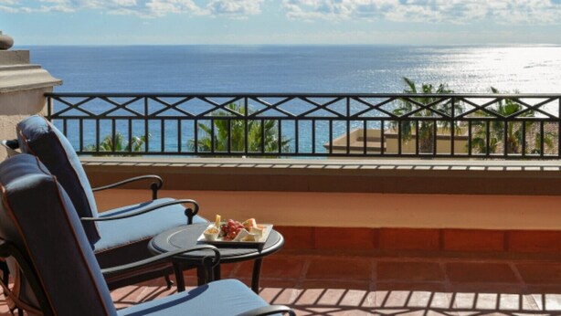 A balcony with a view of the ocean, a table with a plate of food, and two chairs.
