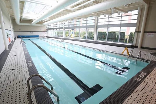Piscina interior com pistas demarcadas, uma cadeira de salva-vidas e uma parede com janelas.