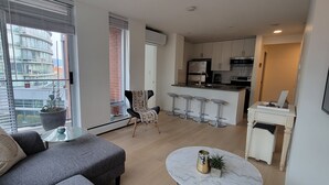Un salon moderne avec un canapé gris, une petite table ronde et une grande fenêtre équipée de stores.