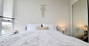 Uma cama bem arrumada com roupa de cama branca, um detalhe em macramé na parede e um abajur ao lado.