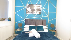 Een slaapkamer met een geometrisch wandontwerp, een bed met een blauwe sprei, twee nachtkastjes met lampen, en een raam met gordijnen.