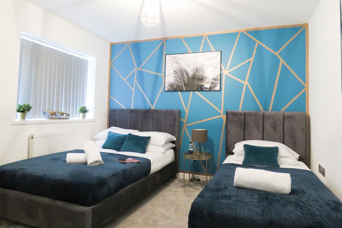A bedroom with two beds, a window with blinds, a wall with a geometric pattern, and a ceiling light.