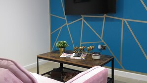 Een kamer met een blauwe geometrische wand, een flatscreen televisie boven een houten salontafel en een roze stoel.