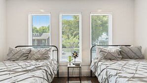 Quarto com duas camas, uma mesinha e três janelas com persianas.