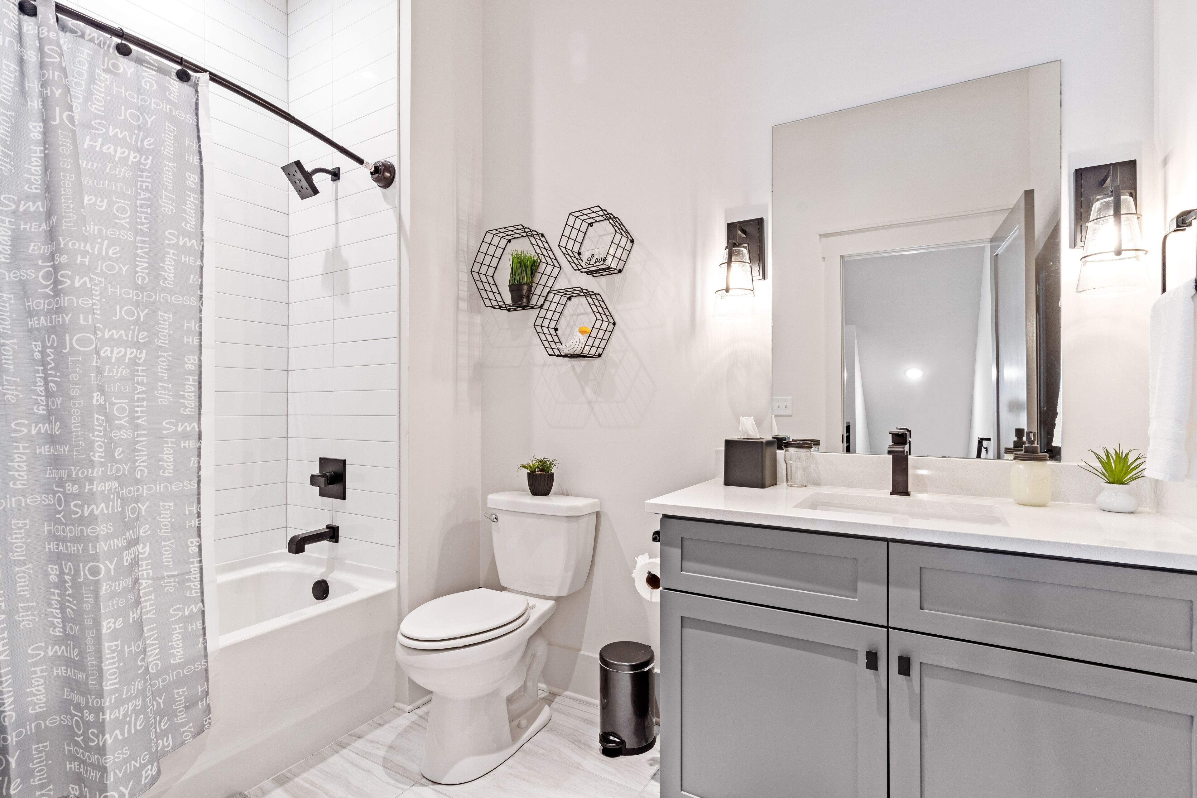 Une salle de bain moderne avec des toilettes blanches, une baignoire, un meuble-lavabo avec un miroir et un rideau de douche orné de mots inspirants.