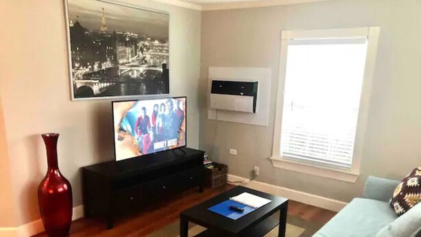 Ein Wohnzimmer mit einem Flachbildfernseher, einer roten Vase, einem schwarzen Couchtisch und einem hellblauen Sofa.