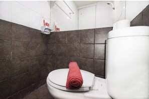 Une salle de bain avec des toilettes blanches, une serviette rouge posée sur le couvercle, et un distributeur de savon fixé au mur.