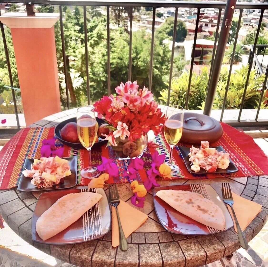 Una mesa dispuesta al aire libre con platos de comida, copas de vino y un jarrón de flores.