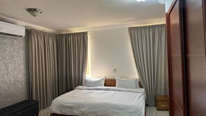 A hotel room with a bed, curtains, a wooden wardrobe, and a wall-mounted air conditioning unit.