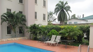 Una piscina con sillas de descanso, rodeada de vegetación y un edificio con múltiples ventanas.