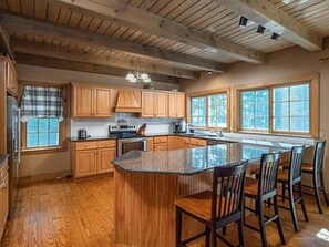 Une cuisine spacieuse avec des meubles en bois, un grand îlot central doté de tabourets de bar et une vue sur l’extérieur grâce à de grandes fenêtres.