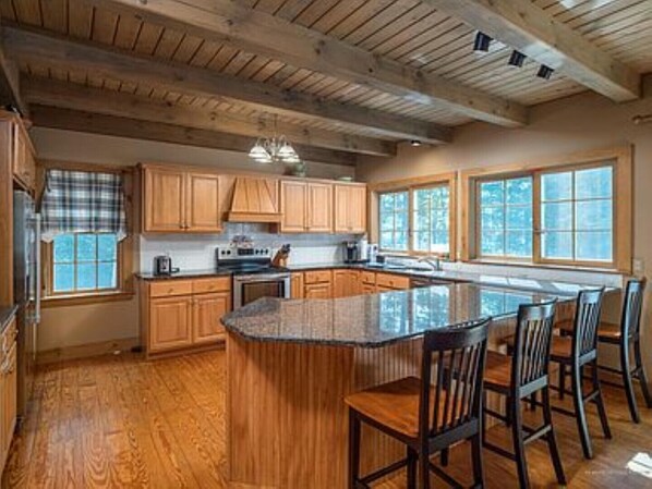 A spacious kitchen with wooden cabinets, a large island with bar stools, and a view of the outdoors through large windows. - Expansive Retreat Near the Slopes w/ Media Room, Two Kitchens, & Stone Fireplace (Newry)