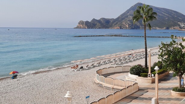 A beach with sun loungers and umbrellas, a palm tree, and a mountain in the background. - Apartment in front of the beach and with free parking in the same building. (Altea)