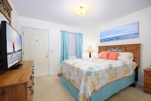 A bedroom with a bed, a television on a wooden dresser, a painting on the wall, and a window with curtains.