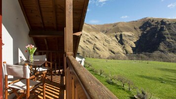 Un balcón de madera con mesa y sillas, con vistas a un campo verde y montañas.