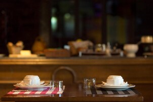 Una mesa de comedor con dos tazas blancas sobre platillos, un posavasos de cuadros y saleros y pimenteros.
