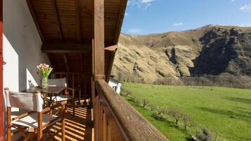 Un balcone in legno con tavolo e sedie, affacciato su un prato verde e montagne.