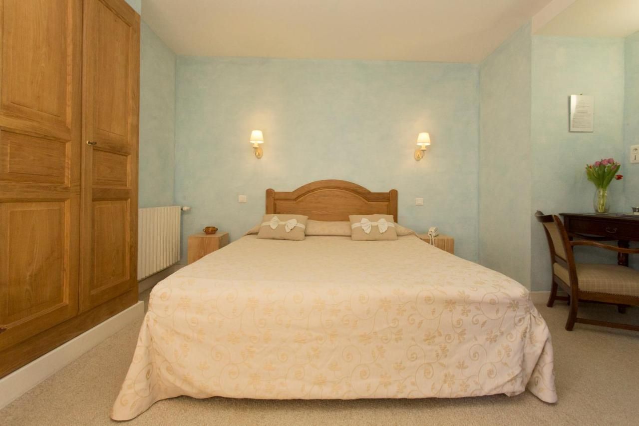 Une chambre avec un grand lit, deux lampes fixées au mur, une armoire en bois, un bureau avec une chaise et un vase rempli de fleurs.