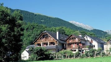 A chalet-style building with a balcony, surrounded by lush greenery and mountains in the background.