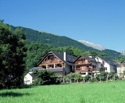 Un edificio de estilo chalet con balcón, rodeado de vegetación exuberante y montañas al fondo.
