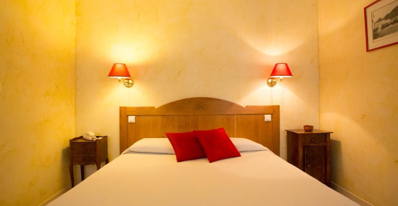 Une chambre d’hôtel avec une tête de lit en bois, deux lampes de chevet, un lit blanc avec des oreillers rouges et une table de chevet sur laquelle se trouvent un téléphone et une petite table.