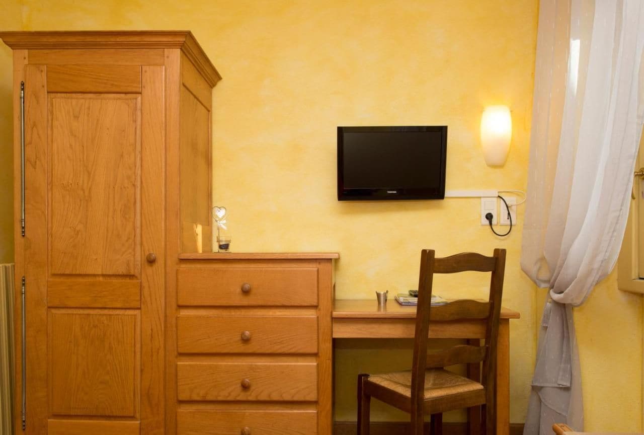 Ein Zimmer mit einem Holzschrank, einem Schreibtisch mit Stuhl, einem an der Wand befestigten Flachbildfernseher und einem Vorhang am Fenster.