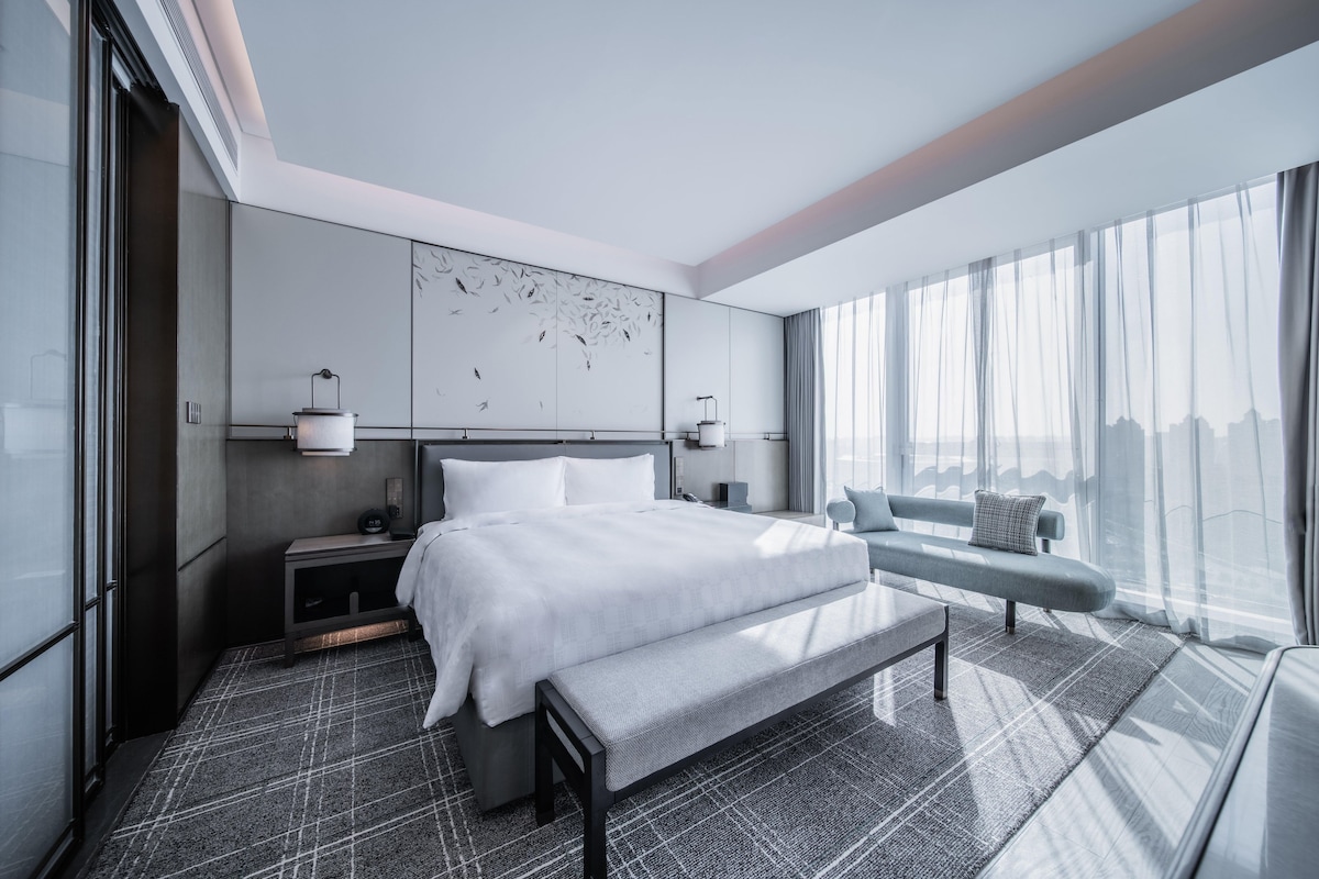 Habitació d'hotel moderna amb un llit gran, una banca, un escriptori i vista a la ciutat.