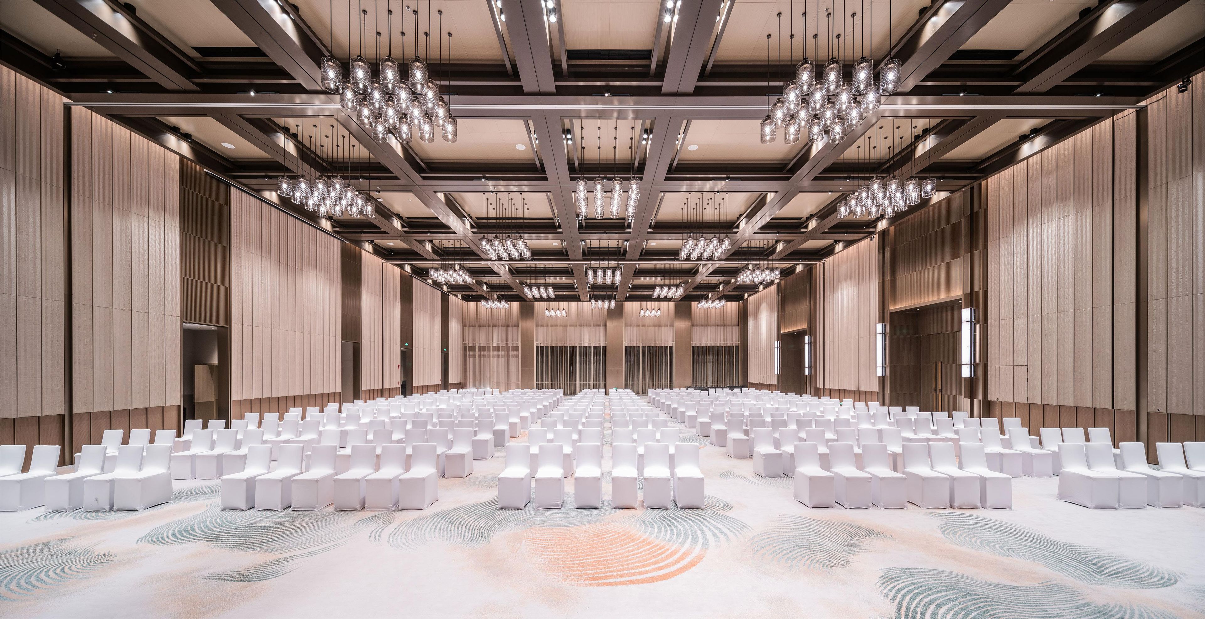 A large, modern conference room with rows of white chairs, a patterned carpet, and a ceiling with hanging light fixtures.