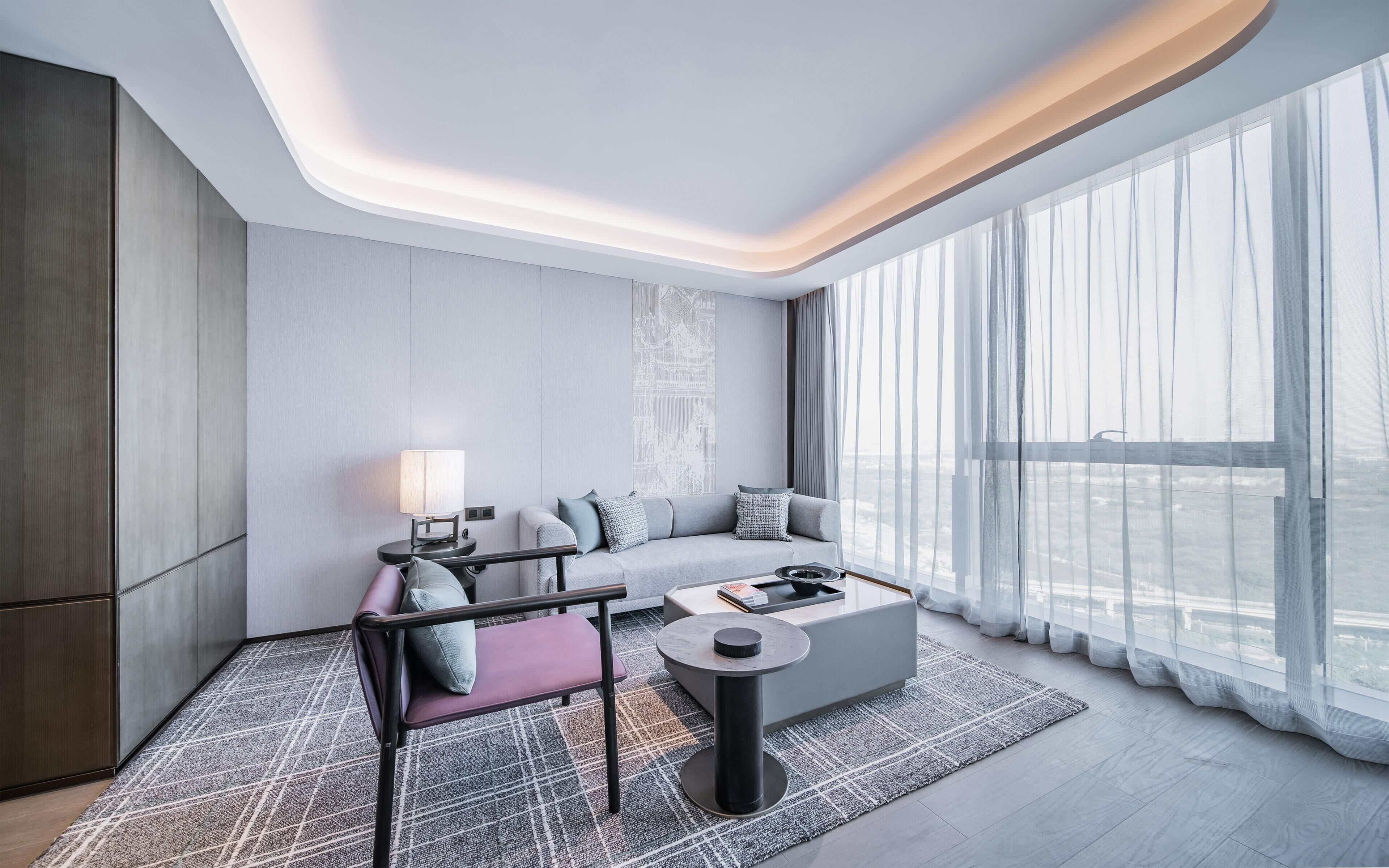 A modern hotel room with a sofa, armchair, and coffee table, large windows with sheer curtains, and a city view.