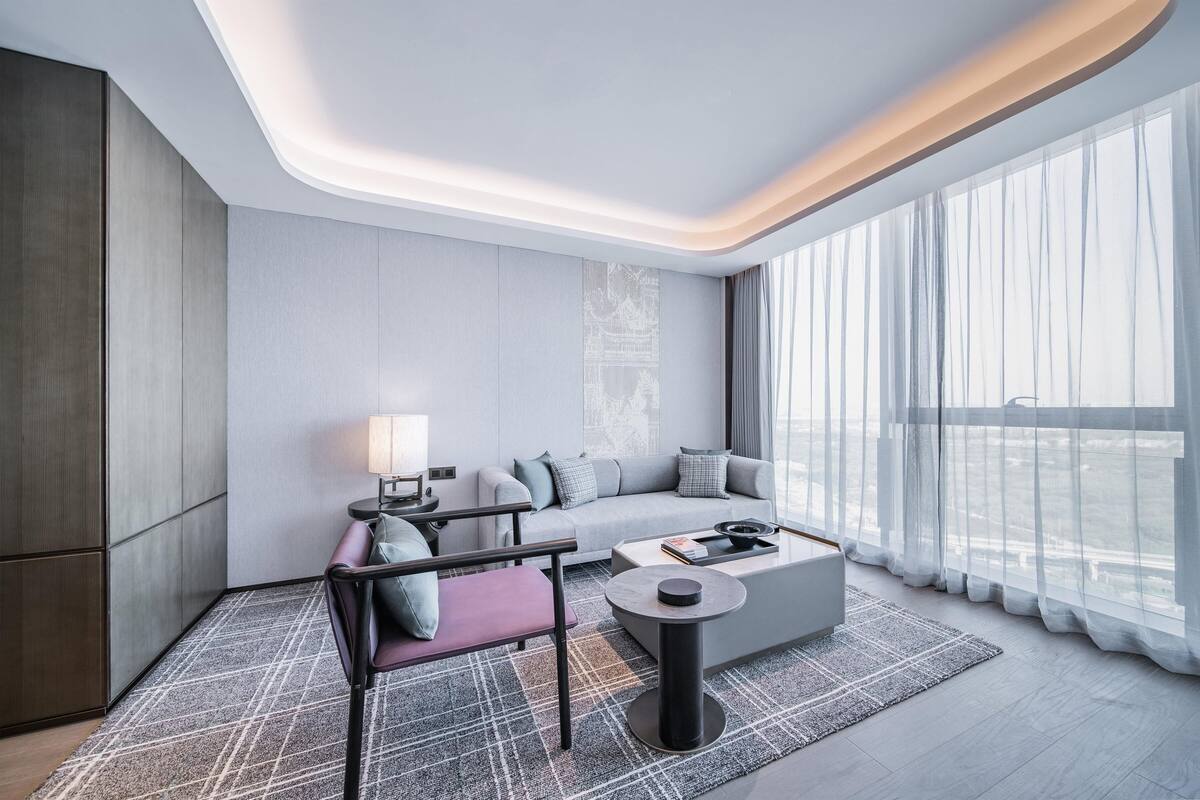 A modern hotel room with a sofa, armchair, and coffee table, large windows with sheer curtains, and a city view.