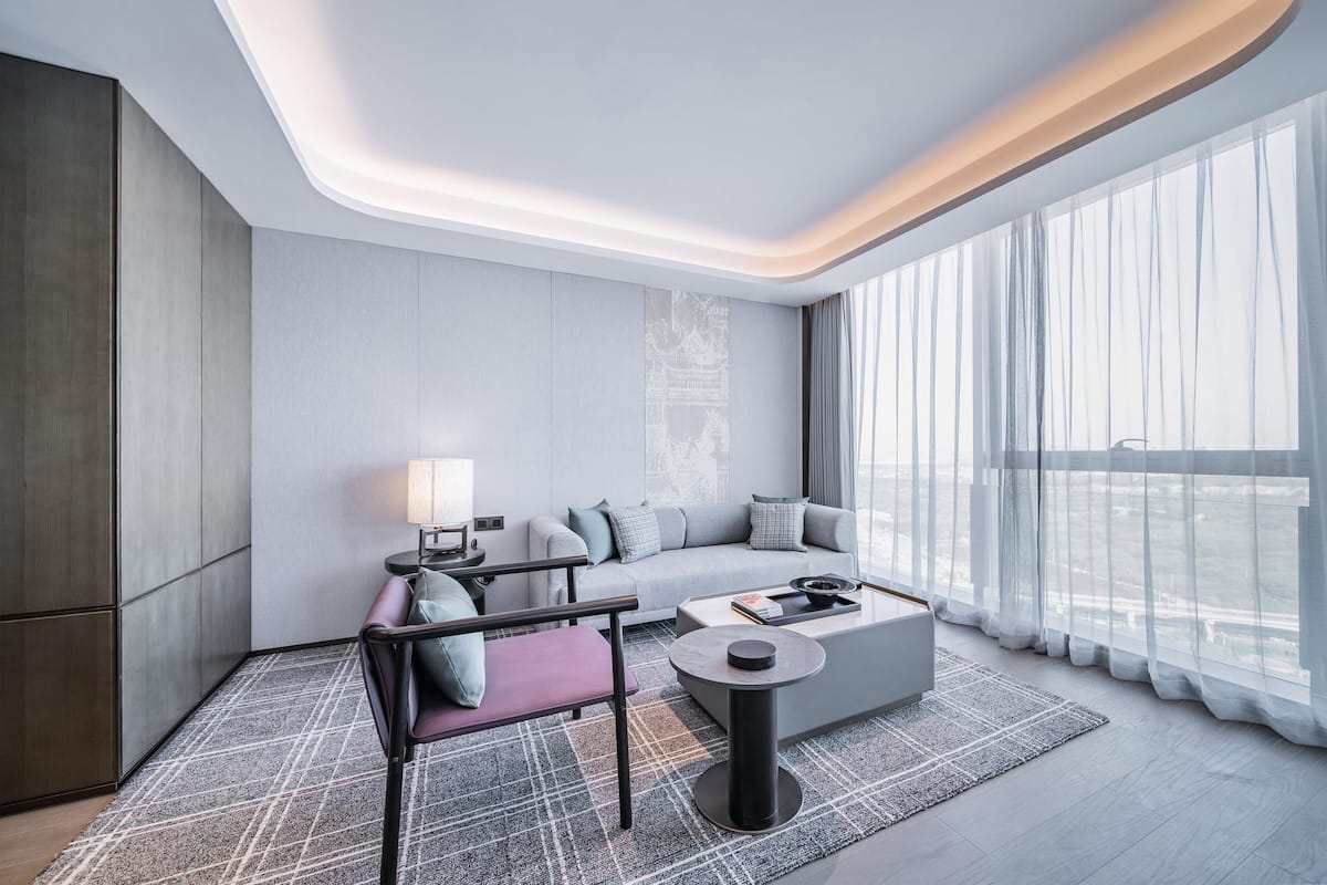 A modern hotel room with a sofa, armchair, and coffee table, large windows with sheer curtains, and a city view.