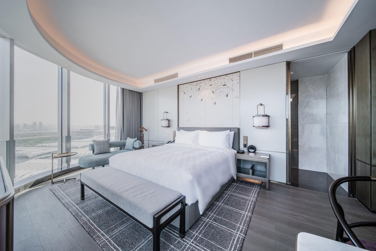 Una habitació d'hotel moderna amb un llit gran, una banca, un escriptori i vistes a la ciutat.