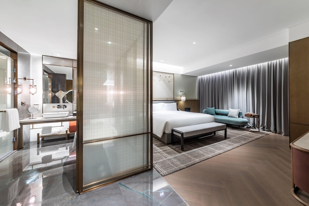 A modern hotel room with a large bed, a sitting area, a bathroom with a vanity, and a large window.