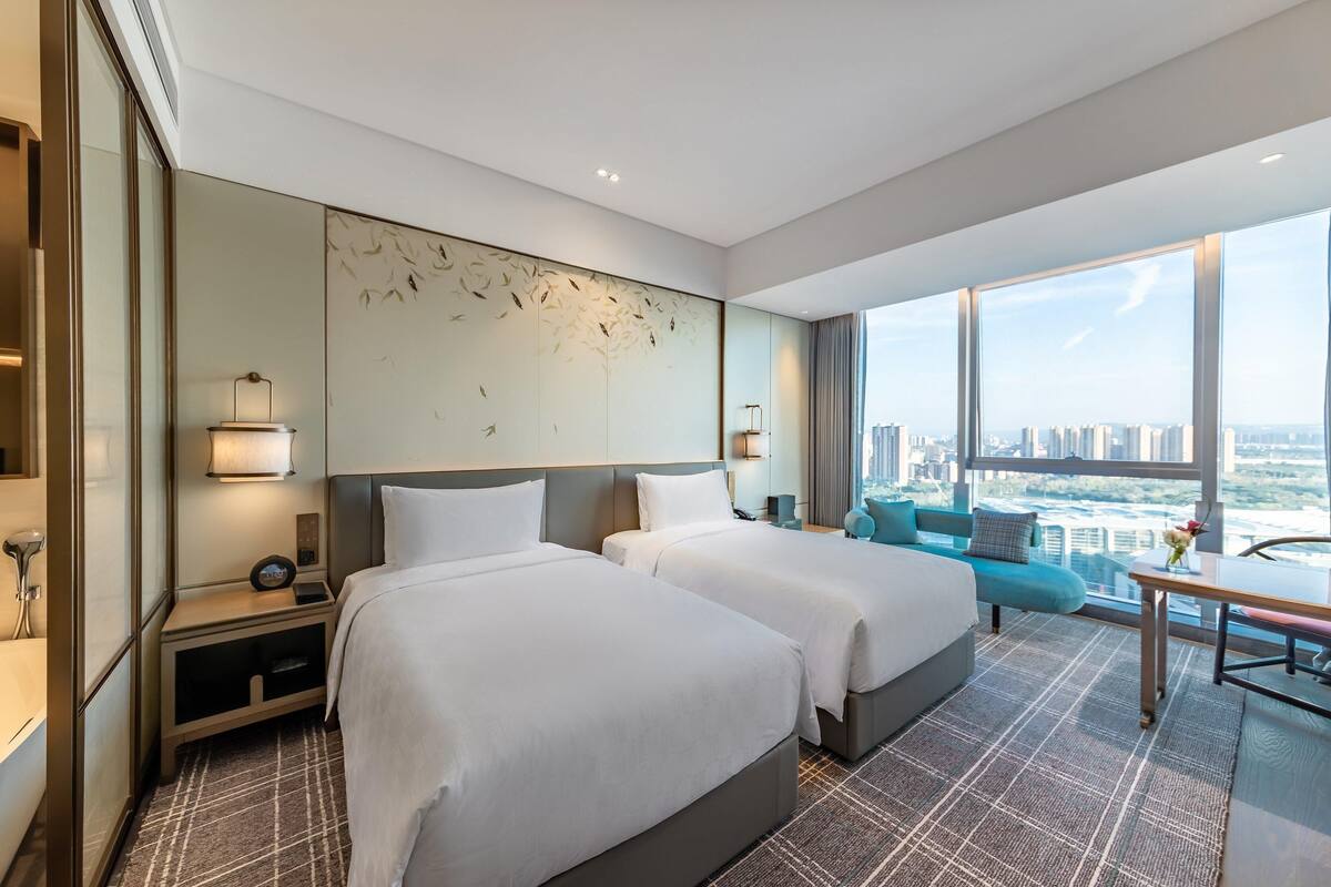 A modern hotel room with two beds, a city view, a large artwork on the wall, and a small table with a vase.